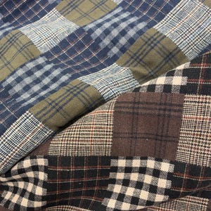 May include: Close-up of patchwork fabric with various plaid patterns. The fabric features squares of blue, olive green, brown, and black and white plaid. The fabric is folded, showing the texture and different patterns.