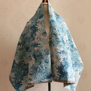 May include: A rectangular textile piece featuring a dense, woven floral pattern in shades of teal, light blue, and cream. The fabric displays a repeating motif of large, stylized chrysanthemum-like blossoms with visible frayed edges along the bottom hem.