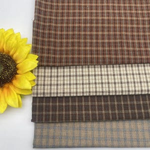 Può includere: Primo piano di quattro campioni di tessuto a quadri in tonalità marroni, beige e blu. Un grande girasole giallo è nell'angolo in alto a sinistra. I tessuti hanno un'estetica classica e rustica, adatta a vari progetti di artigianato e decorazione.