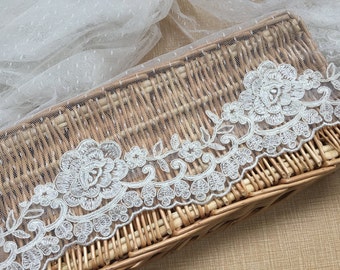 May include: White lace trim featuring a floral design. The lace is delicate and intricate, with small flowers and leaves. The lace is displayed on a wicker basket.