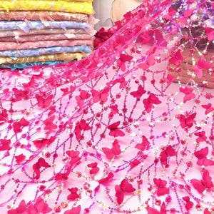 May include: Pink tulle fabric with 3D butterfly and sequin embellishments. The butterflies are a vibrant fuchsia color, and the sequins add a touch of sparkle. The fabric is layered over a stack of colorful fabrics.