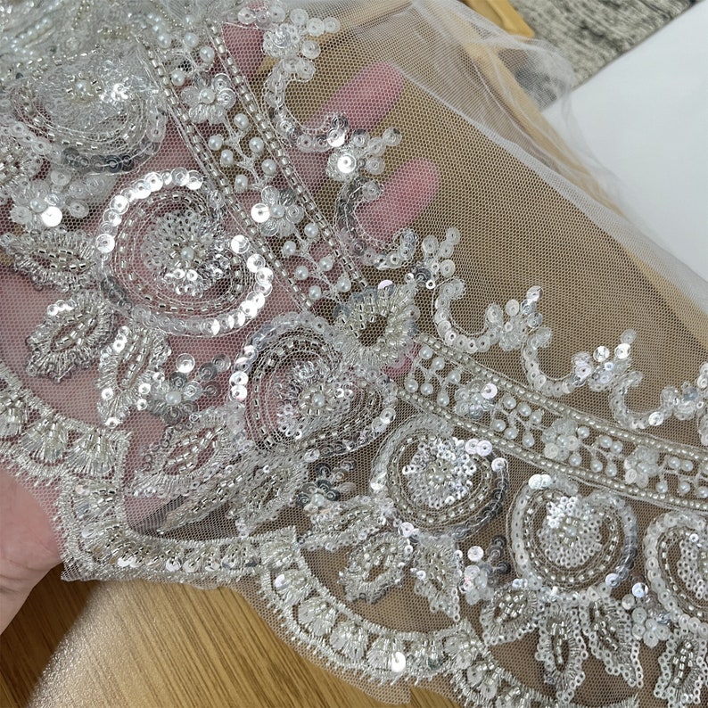 Pu&ograve; includere: Bordino in pizzo argentato e bianco con perline e paillettes con un retro in rete trasparente.