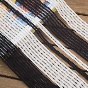 Peut inclure: Plusieurs bandes de galon décoratif présentant un motif géométrique répétitif de lignes verticales et de petits carrés ajourés. Le galon est disponible en noir uni et en blanc uni, chaque bande affichant une texture uniforme et cohérente.