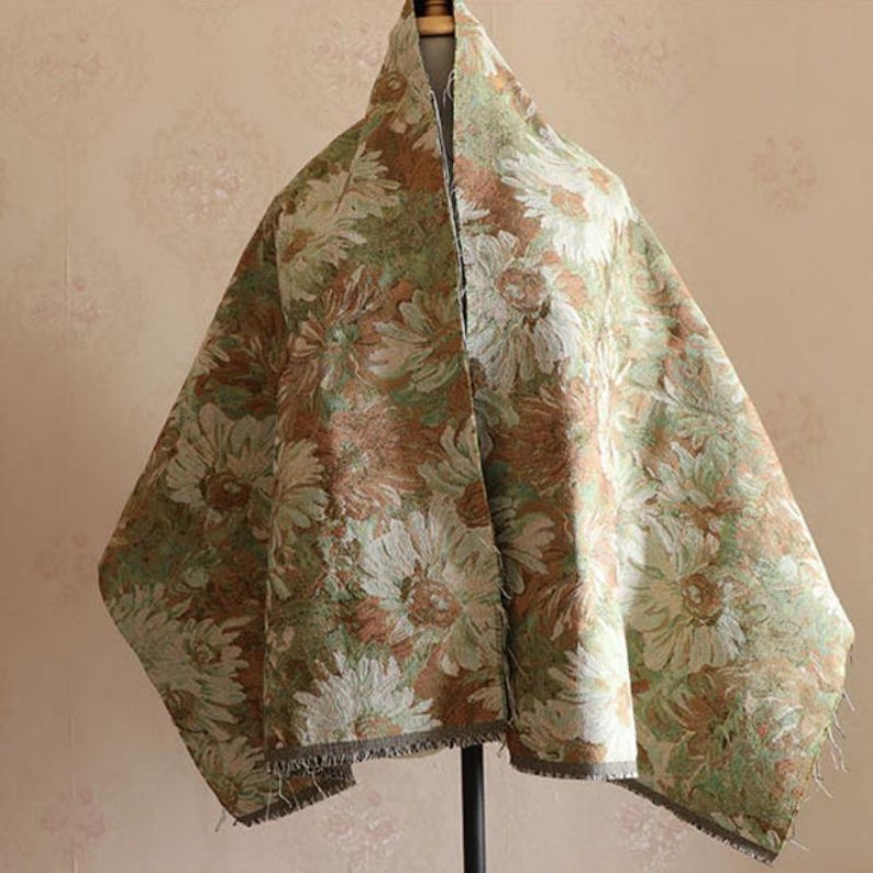 May include: A rectangular fabric piece featuring a dense, woven floral pattern in shades of sage green, muted brown, and cream. The textile displays large, overlapping daisy-like flowers across the entire surface, finished with a raw, frayed edge.