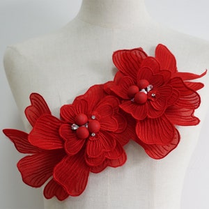 May include: Two red fabric flowers with layered petals and rhinestone accents. The flowers are attached to a white background.