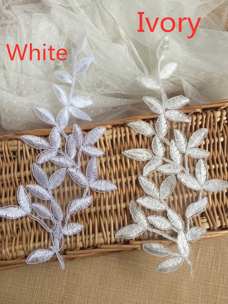 May include: Two white lace appliques in the shape of leafy branches. The appliques are on a woven wicker surface. The text "White" and "Ivory" is visible above the appliques.