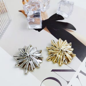 May include: Two decorative starburst-shaped objects, one silver and one gold, are displayed on a white surface. The objects have a textured, ridged design. Clear ice cubes are scattered around the objects.