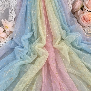May include: A sheer, multi-colored tulle fabric featuring a gradient of pastel blue, yellow, and pink hues. The lightweight, semi-transparent material is decorated with scattered, shimmering gold foil flecks throughout the entire surface.