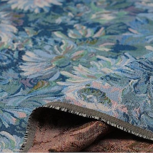 May include: A woven fabric featuring a dense floral pattern in shades of blue, teal, and light green. The textile displays a textured surface with visible threads and a folded edge revealing a contrasting dark brown underside with a woven design.
