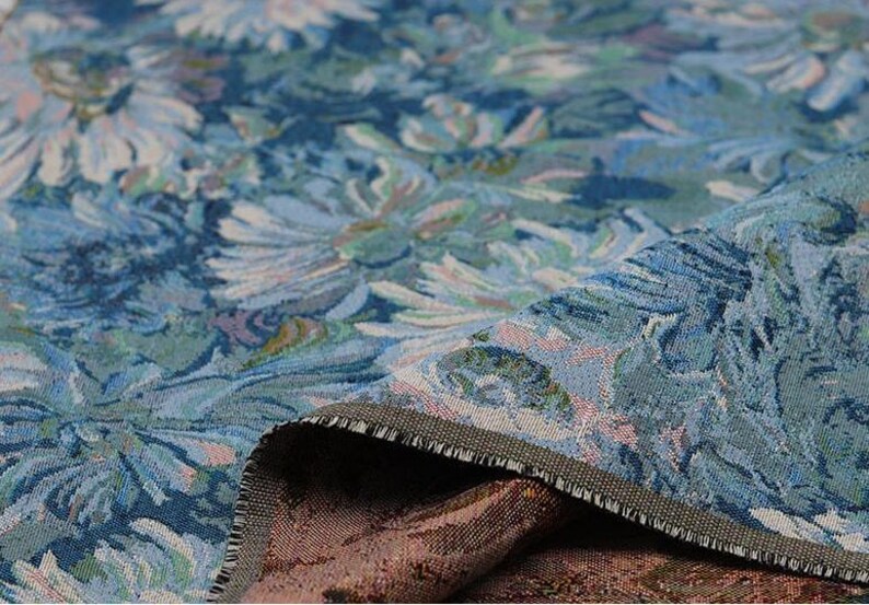 May include: A woven fabric featuring a dense floral pattern in shades of blue, teal, and light green. The textile displays a textured surface with visible threads and a folded edge revealing a contrasting dark brown underside with a woven design.