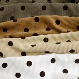 May include: Four layers of corduroy fabric stacked horizontally, each featuring a repeating pattern of dark brown polka dots. The colors of the fabric layers from top to bottom are dark olive green, golden tan, light beige, and crisp white.