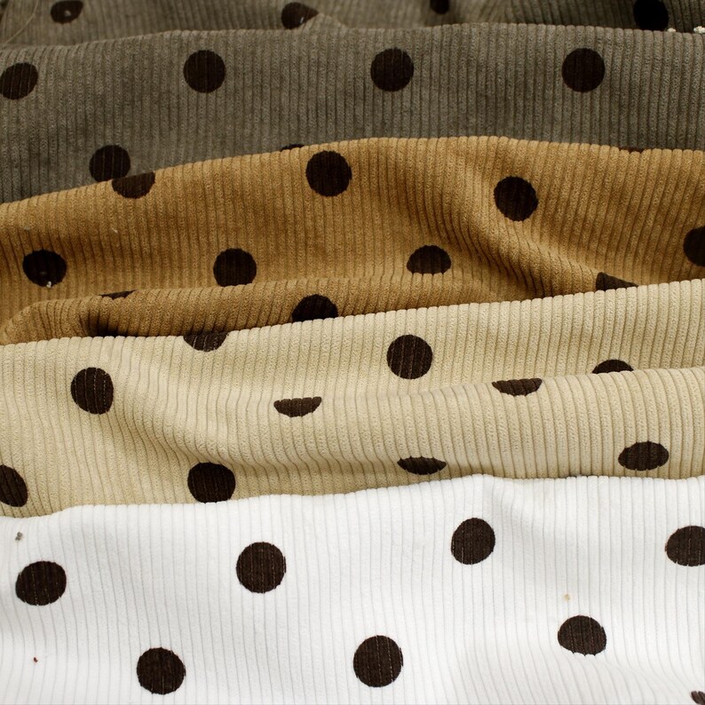 May include: Four layers of corduroy fabric stacked horizontally, each featuring a repeating pattern of dark brown polka dots. The colors of the fabric layers from top to bottom are dark olive green, golden tan, light beige, and crisp white.