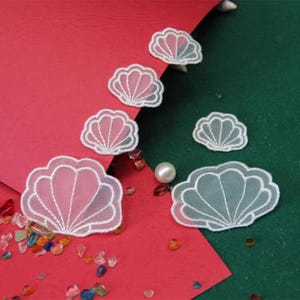 Pode incluir: Apliques brancos em forma de concha com um design delicado de renda. Os apliques estão dispostos sobre uma superfície vermelha e verde, com uma dispersão de pedras preciosas coloridas e uma única pérola. As conchas têm um interior rosa.