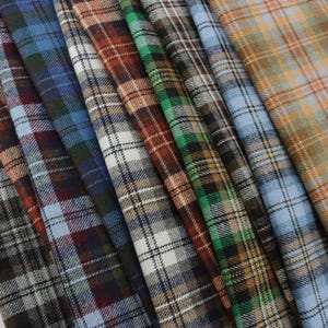 May include: A selection of plaid fabric samples in shades of blue, brown, green, and beige. The fabric features a traditional checked design, with each piece folded and arranged to showcase the diverse colour schemes and textures.