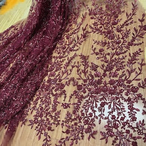 Peut inclure: Tissu en dentelle bordeaux avec des motifs floraux et de feuilles. Le tissu est orné de paillettes, ajoutant un effet scintillant. Le design présente des éléments botaniques détaillés, créant un textile luxueux et élégant.