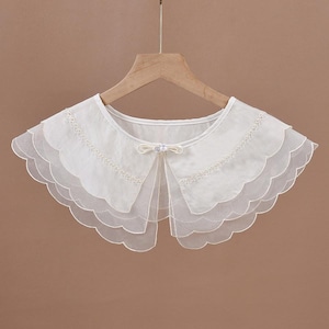 May include: A white detachable collar featuring three layers of scalloped sheer fabric. The collar is adorned with delicate floral embroidery along the top edge and is secured at the center front with a small ribbon bow and a single white pearl bead.