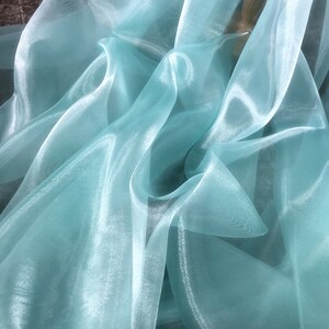 K&ouml;nnte beinhalten: Nahaufnahme eines hellblauen, durchsichtigen Stoffes mit glatter, seidenartiger Textur. Der Stoff ist drapiert und gefaltet und erzeugt weiche, flie&szlig;ende Linien.