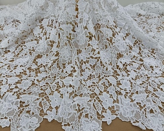 May include: A white lace fabric featuring an intricate floral pattern with detailed petals and leaves. The design consists of interconnected blossoms and foliage, creating a delicate, openwork texture throughout the entire piece of textile.