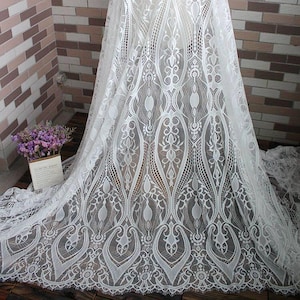 May include: White lace fabric with an intricate floral and geometric pattern. The delicate material is draped, showcasing the detailed openwork design. The fabric is suitable for bridal wear, dresses, or decorative projects.