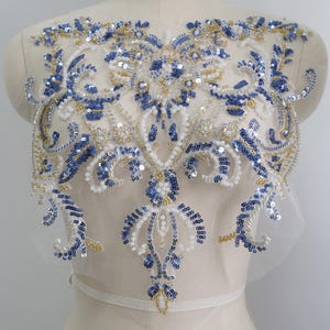 Puede incluir: Aplicación bordada ornamentada con un diseño simétrico. Presenta lentejuelas azules, cuentas blancas y detalles dorados. La aplicación está colocada sobre una tela transparente blanquecina, creando un elemento decorativo delicado. Ideal para adornar prendas o accesorios.