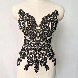 Peut inclure: Une application en dentelle noire avec des motifs tourbillonnants complexes et des perles denses, présentée sur un buste de mannequin beige. Le motif couvre la poitrine et le milieu du corps avec un ornement symétrique en broderie texturée.