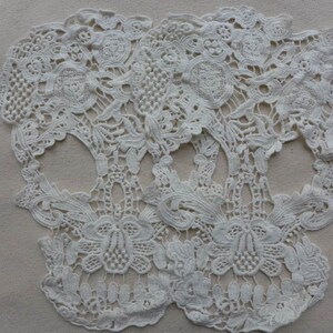 May include: Two white lace skull shapes, with intricate floral designs, are placed on a beige fabric background.