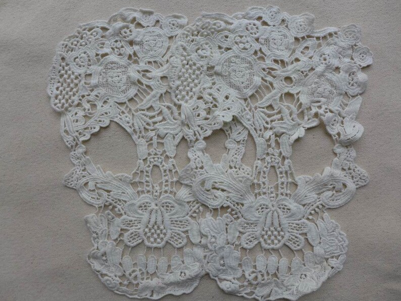May include: Two white lace skull shapes, with intricate floral designs, are placed on a beige fabric background.