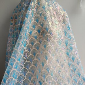 May include: A sheer fabric featuring a repeating scalloped pattern embroidered with iridescent blue and white sequins. The design creates a fish scale effect with shimmering light blue and pearlescent accents throughout the translucent material.