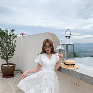 Puede incluir: Un vestido blanco de encaje con escote en V y mangas cortas. El vestido tiene un corpi&ntilde;o ajustado y una falda acampanada. El vestido est&aacute; decorado con un delicado patr&oacute;n de encaje.