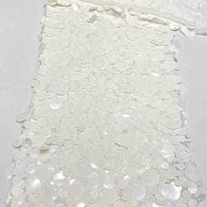 Puede incluir: Tela color blanco roto con una capa densa de lentejuelas circulares superpuestas de varios tama&ntilde;os. Las lentejuelas est&aacute;n fijadas sobre una base transparente, creando una superficie texturizada y reflectante de color uniforme.
