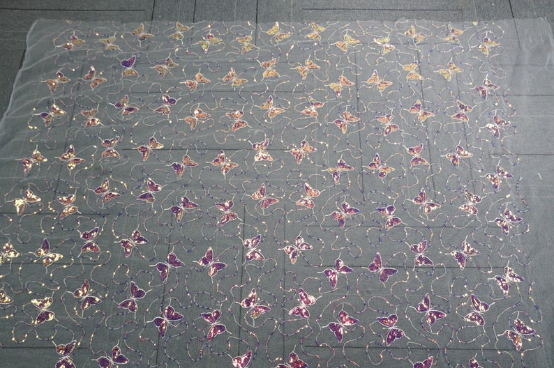 May include: A sheer fabric with a repeating pattern of purple and gold butterfly sequins. The fabric is white and has a subtle shimmer.