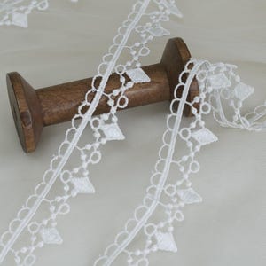 May include: White lace trim with a delicate diamond and scalloped edge design. The lace is spooled on a wooden bobbin. This trim is ideal for sewing, crafting, and adding a decorative touch to various projects.