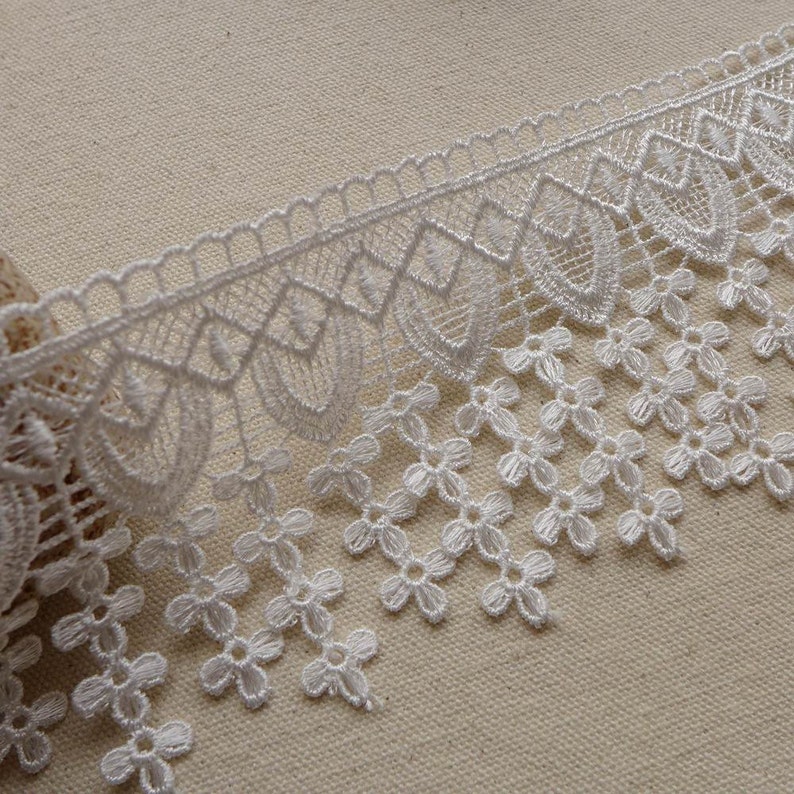 May include: White lace trim with a scalloped edge and a fringe of small flowers. The lace is delicate and intricate, with a floral pattern.