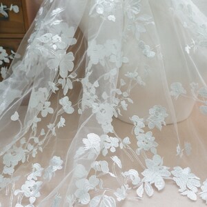 May include: White floral lace fabric with a delicate pattern of flowers and leaves. The lace is sheer and has a soft, flowing texture.