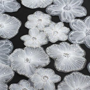 Może przedstawiać: Zbliżenie na kilka białych, przezroczystych aplikacji kwiatowych z tkaniny. Każdy kwiat ma perełkowe centrum i obramowanie z cekinami. Płatki są warstwowe, tworząc efekt trójwymiarowy. Kwiaty są rozrzucone na czarnym tle.