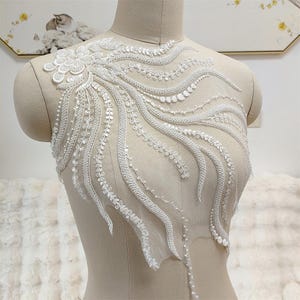May include: A white lace appliqué featuring intricate floral and swirling vine patterns embellished with small beads and sequins. The design is arranged on a sheer mesh base, showcasing a dense cluster of petals at the top with flowing, elongated tendrils extending downwards across the bodice area.