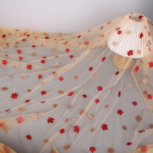 Puede incluir: Una tela de tul beige transparente con un patrón bordado de hojas de arce dispersas en tonos rojo vibrante y dorado brillante. La tela está drapeada sobre una pantalla de lámpara blanca plisada con una base de madera.