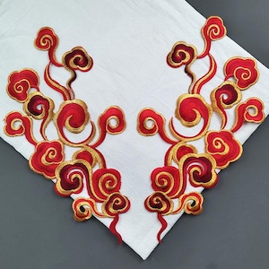 Op de afbeelding: Twee geborduurde patches met een wolkenontwerp. De patches hebben een rood en goud kleurenschema, met wervelende wolkenvormen en ingewikkelde details. Ze zijn geplaatst op een witte stoffen achtergrond.