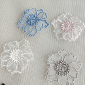 May include: Four floral appliques featuring sheer, translucent fabric petals with embroidered edges and centers. The set includes one blue, one light pink, one white, and one grey flower, each with a detailed, circular stitched stamen design.