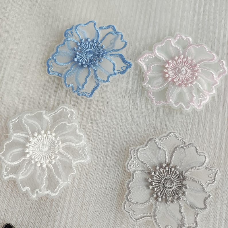 May include: Four floral appliques featuring sheer, translucent fabric petals with embroidered edges and centers. The set includes one blue, one light pink, one white, and one grey flower, each with a detailed, circular stitched stamen design.
