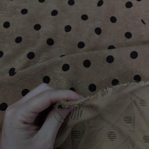 May include: A tan corduroy fabric featuring a repeating pattern of black polka dots. The underside of the fabric is visible, revealing a lighter tan color with a subtle pattern of small, dark, horizontal dashed lines arranged in a grid.