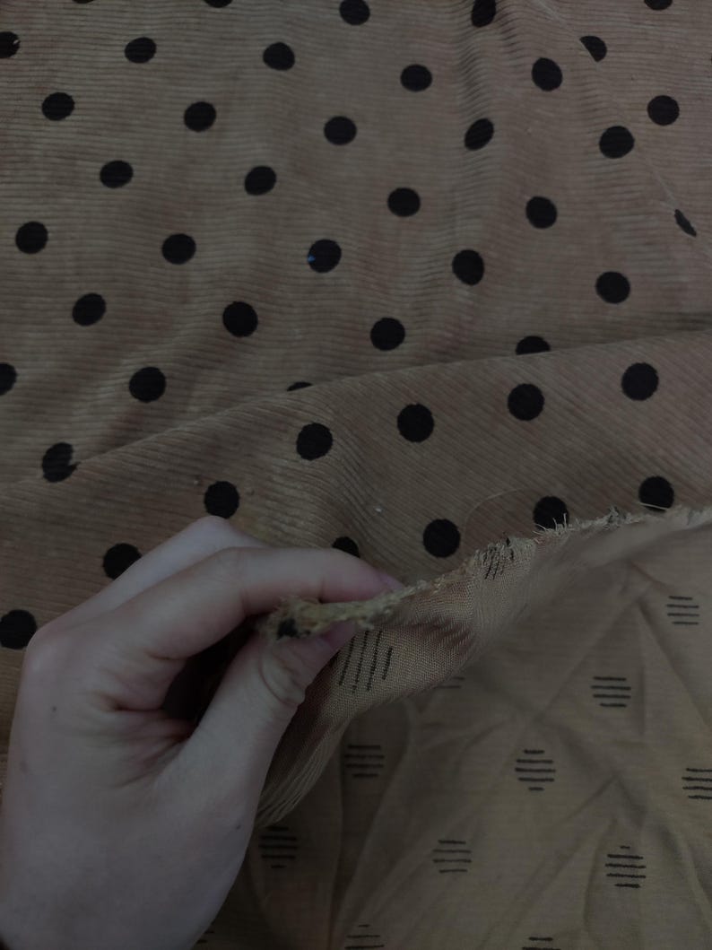 May include: A tan corduroy fabric featuring a repeating pattern of black polka dots. The underside of the fabric is visible, revealing a lighter tan color with a subtle pattern of small, dark, horizontal dashed lines arranged in a grid.