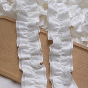May include: Close-up of white ruffled fabric trim. The trim has a wavy, pleated design with a stitched edge. The fabric appears to be lightweight and delicate, suitable for various craft projects. The background is a light wooden surface.