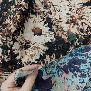 May include: A woven fabric featuring a dense floral pattern with large cream-colored daisy-like flowers set against a dark black background. The textile displays a textured, tapestry-style weave with visible threads in shades of brown, green, and tan.