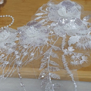 Peut inclure: Applique de dentelle florale blanche avec des paillettes. L'applique présente de multiples fleurs et feuilles, avec des détails complexes et des embellissements scintillants. Idéal pour les vêtements de mariée ou autres projets décoratifs.