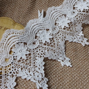 May include: White lace trim with a scalloped edge and floral design. The lace is delicate and intricate, with a repeating pattern of flowers and leaves.
