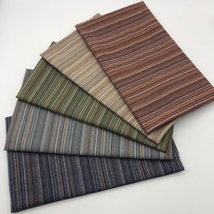Op de afbeelding: Een verzameling van vijf rechthoekige stofstalen die in een waaier zijn gerangschikt. Elk stuk stof heeft een eigen kleurenpalet van fijne verticale strepen, waaronder tinten roest, beige, olijfgroen, leiblauw en donker antracietgrijs.