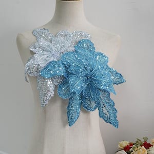 Puede incluir: Dos apliques florales, uno blanco y otro azul claro, se exhiben en un maniquí. Los apliques están muy adornados con lentejuelas y cuentas, creando un efecto brillante. El aplique azul claro está en primer plano, con el blanco detrás.