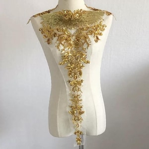 Puede incluir: Una aplicación bordada con lentejuelas y cuentas doradas que presenta un patrón denso y complejo de flores y enredaderas. El diseño se extiende desde una pieza de cuello ancha en forma de abanico hasta una sección vertical larga y estrecha.