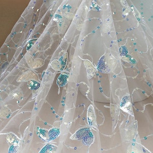 May include: White tulle fabric with iridescent blue and silver butterfly sequins. The fabric is sheer and delicate, with a soft, flowing texture.